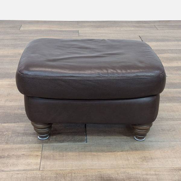 Brown leather ottoman with turned wood bun feet, seen at an angle on wooden flooring.