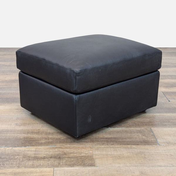 1. Reperch black leather ottoman with clean lines displayed on wooden floor, three-quarter angled view.