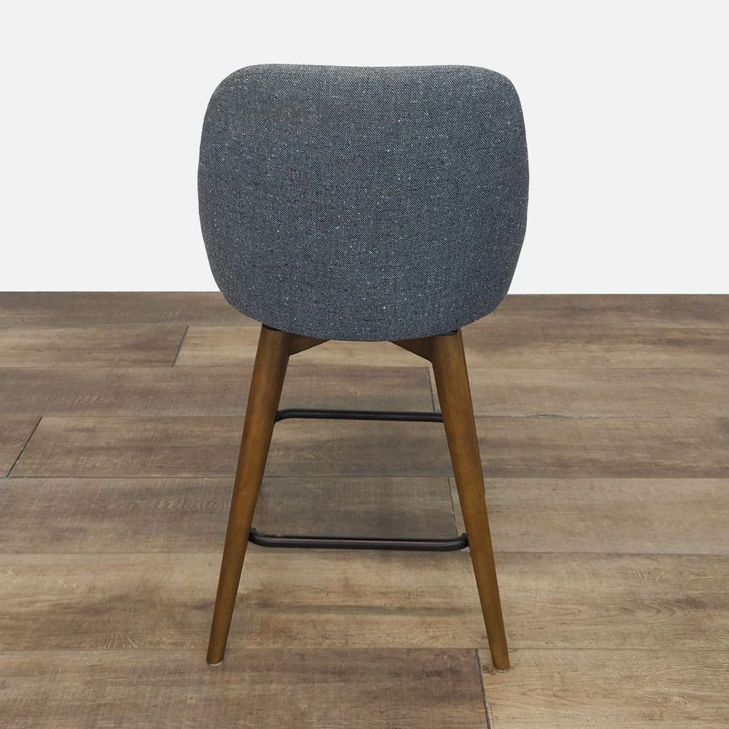 Back view of a gray upholstered stool featuring wood legs and a metal footrest.