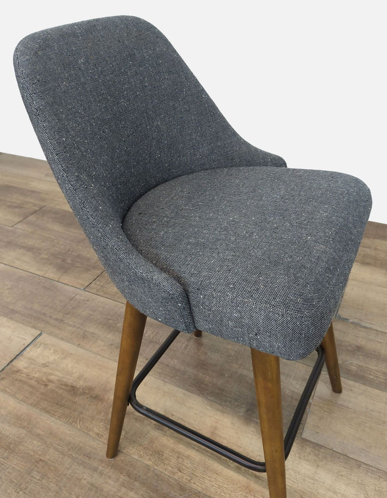Angled view of a gray upholstered stool with wooden legs and metal footrest.