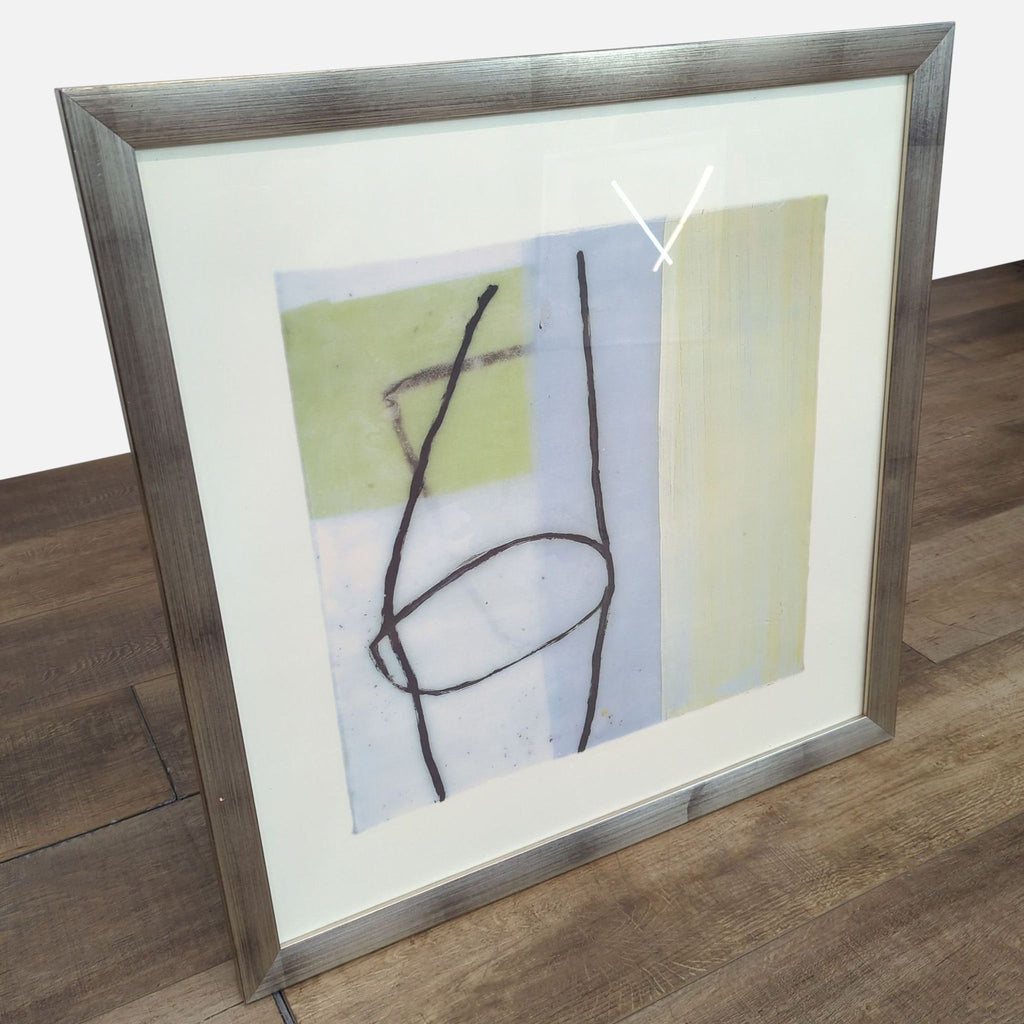 Framed abstract art with geometric elements and silver border, set on wooden flooring.