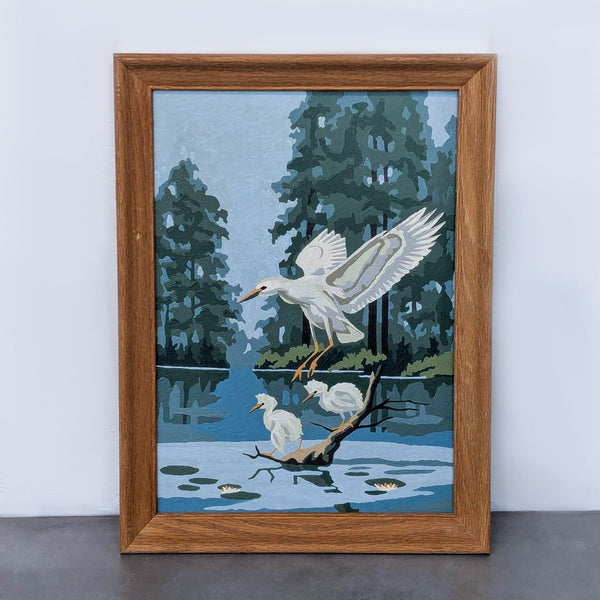Vintage paint by number of flying heron in wood frame, set against trees and water.