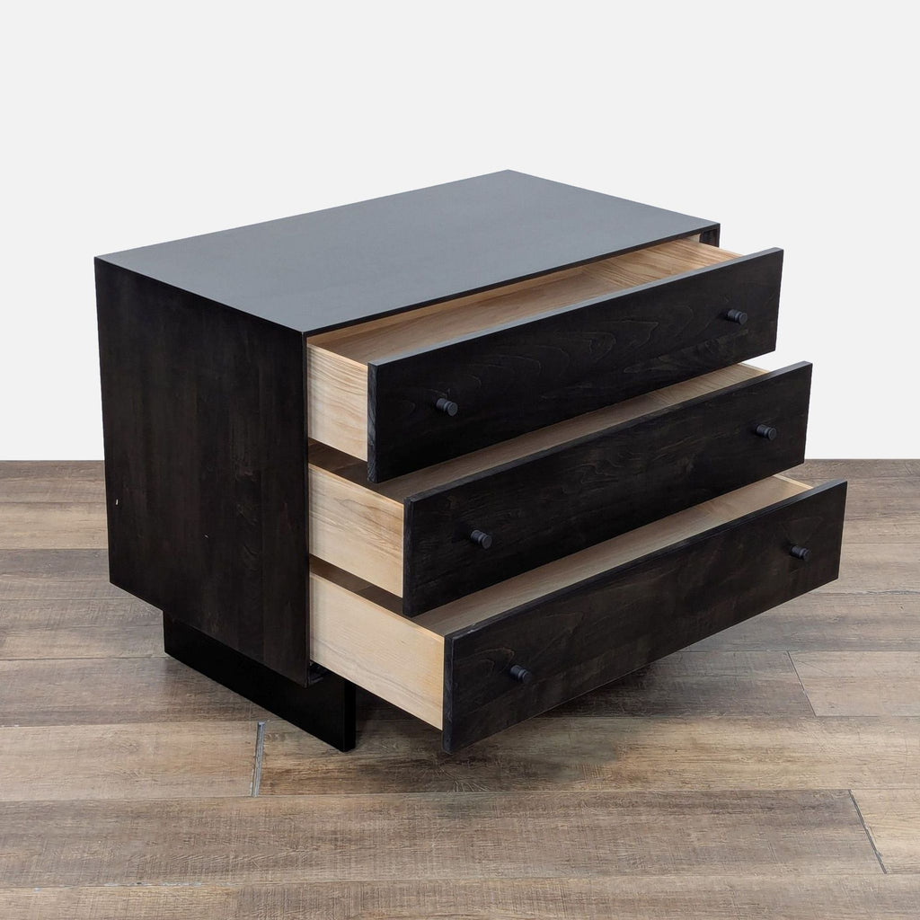Side view of a black wood three-drawer dresser on a wooden floor.