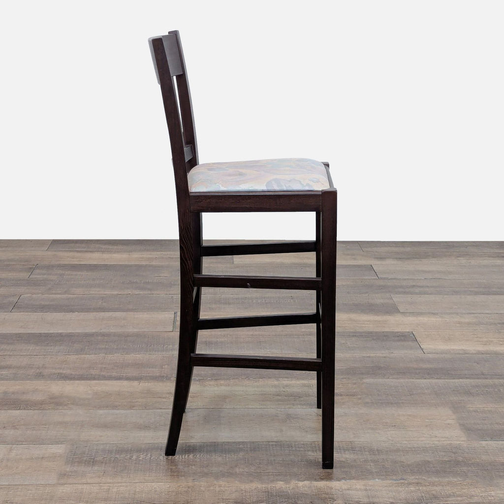 Side view of a dark wooden bar stool with a cushioned seat, placed on wooden flooring.