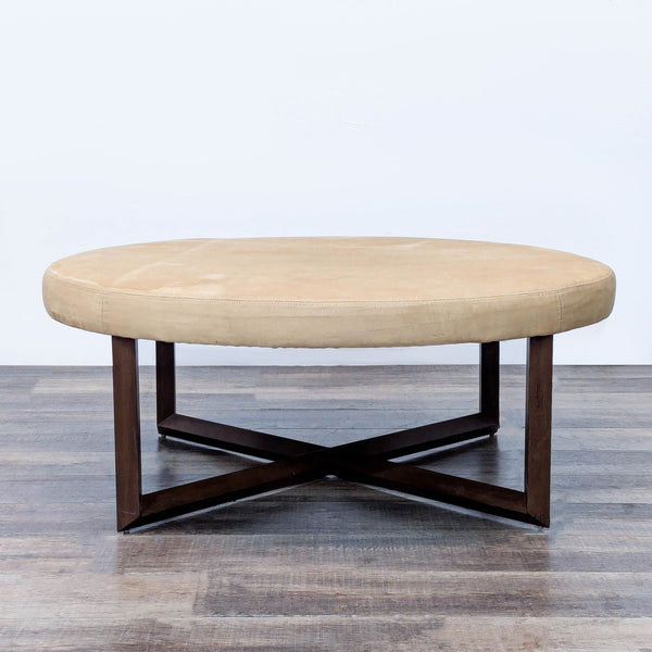 Round beige ottoman with a dark wood cross base on hardwood floor.