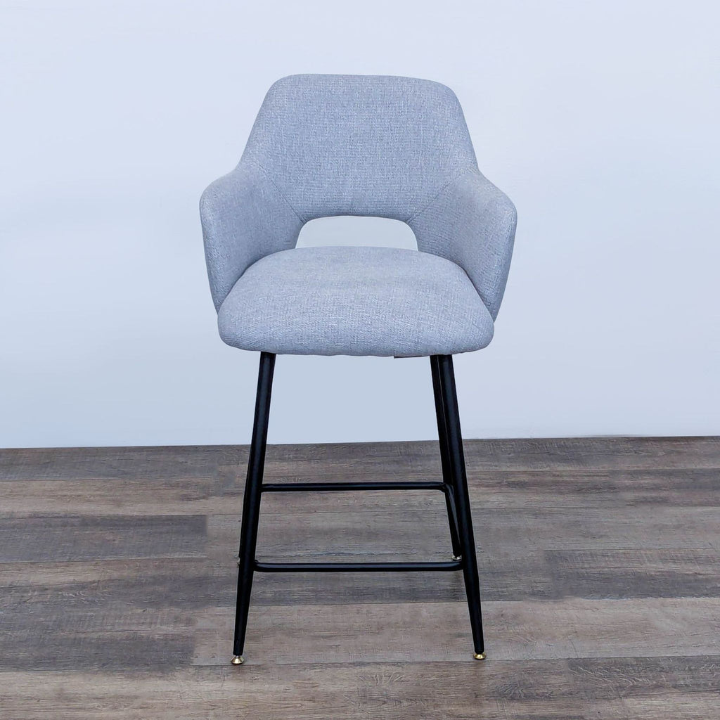 Gray upholstered bar stool with curved backrest and black metal legs on wooden floor.