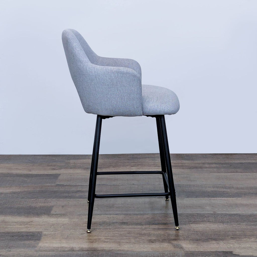 Angled view of gray bar stool by Reperch, with cushioned seat and backrest, metal legs on wood floor.