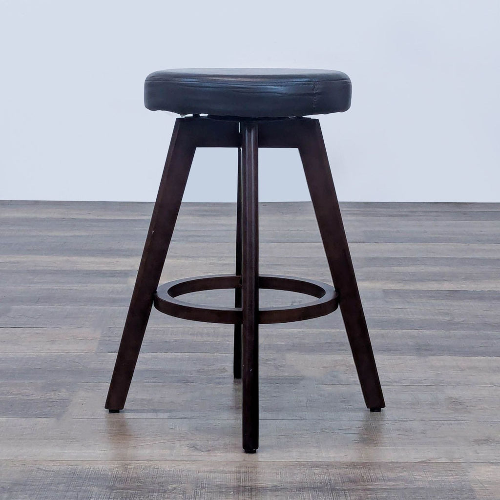 Round black cushioned stool with dark wooden legs and ring base on wood floor.