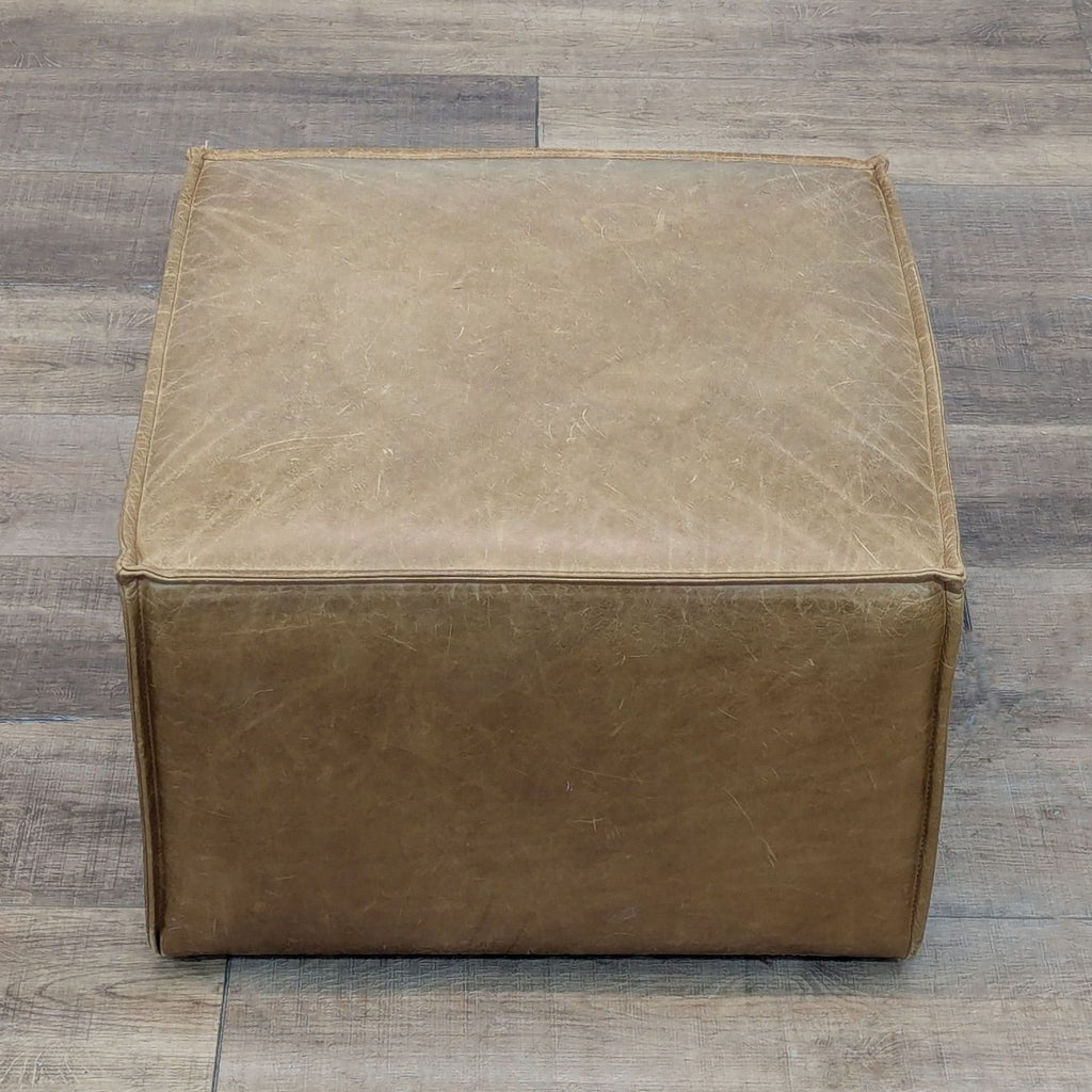 Tan leather cube ottoman with visible seams on wooden floor.