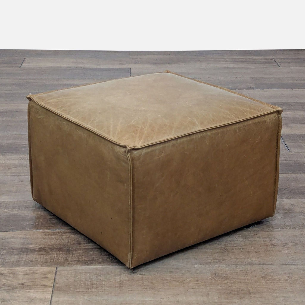 Square leather ottoman in tan, set on wooden flooring.