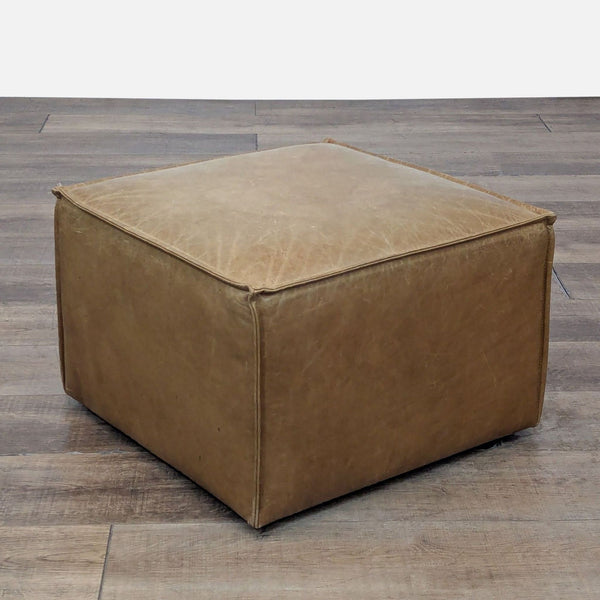 Square leather ottoman in tan, set on wooden flooring.