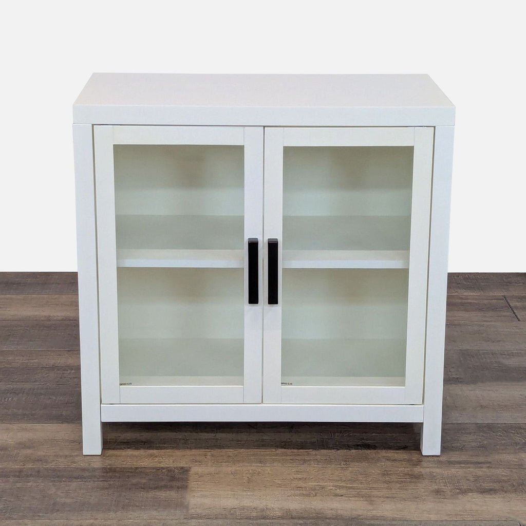 Glass door cabinet with a white frame and black handles on wood floor.
