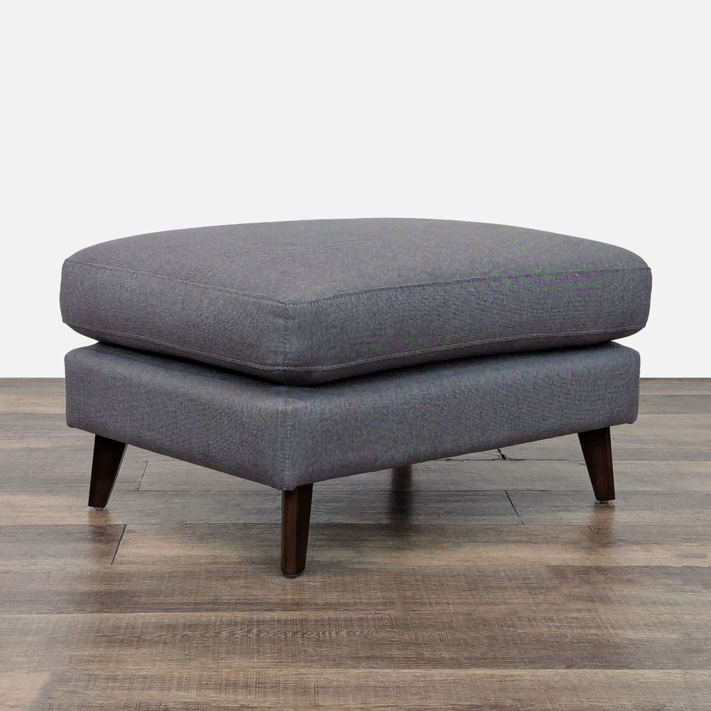 Angled view of a gray ottoman featuring a cushioned top and wooden legs.