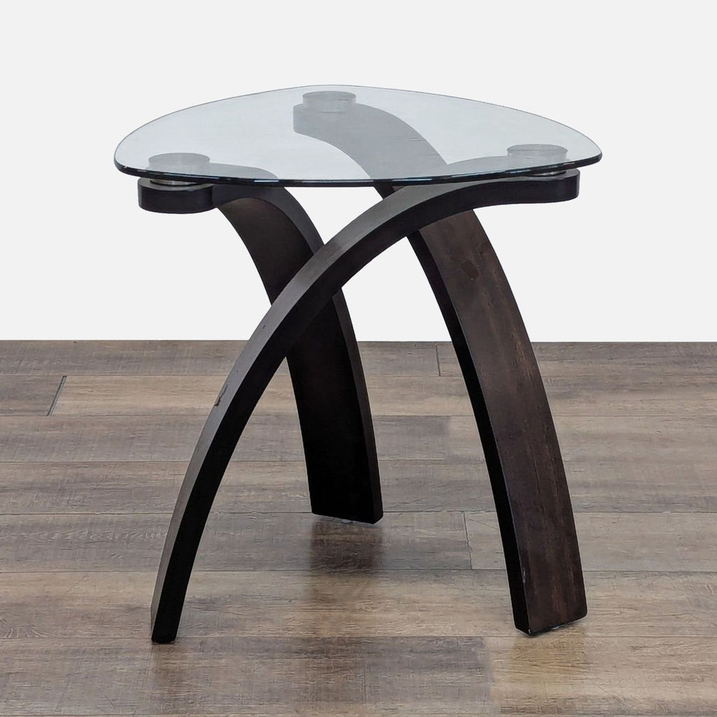 Triangular glass top table with curved hardwood base on wood floor.