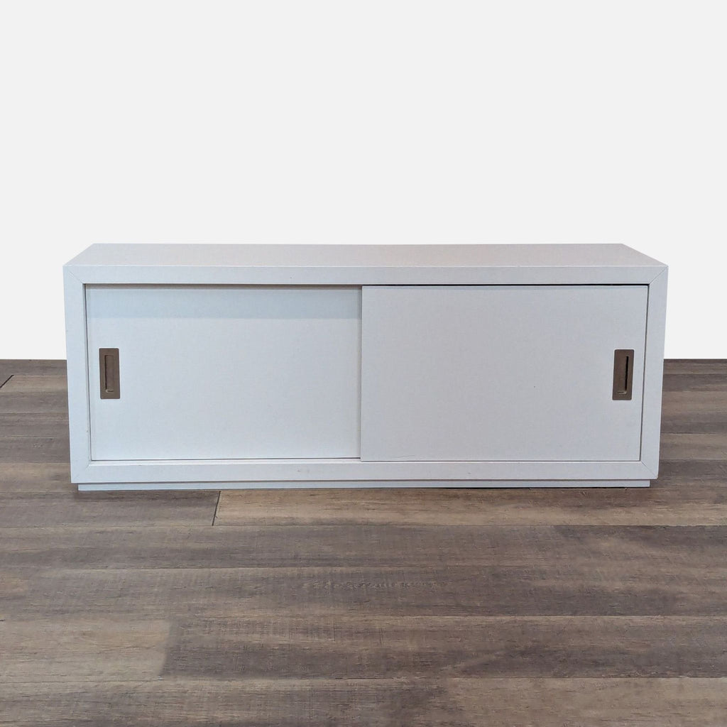 White rectangular wooden cabinet with sliding doors and recessed handles on a wooden floor.  