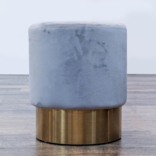 Gray cylindrical stool with a soft velvet top and brass metal base on wooden floor.