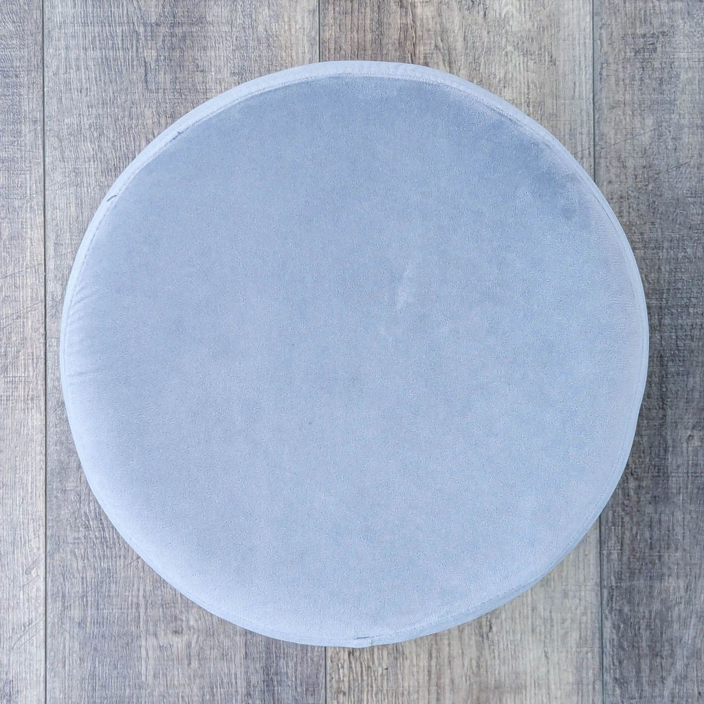Side angle of a gray velvet stool with a cylindrical shape and brass base on a textured wooden floor.