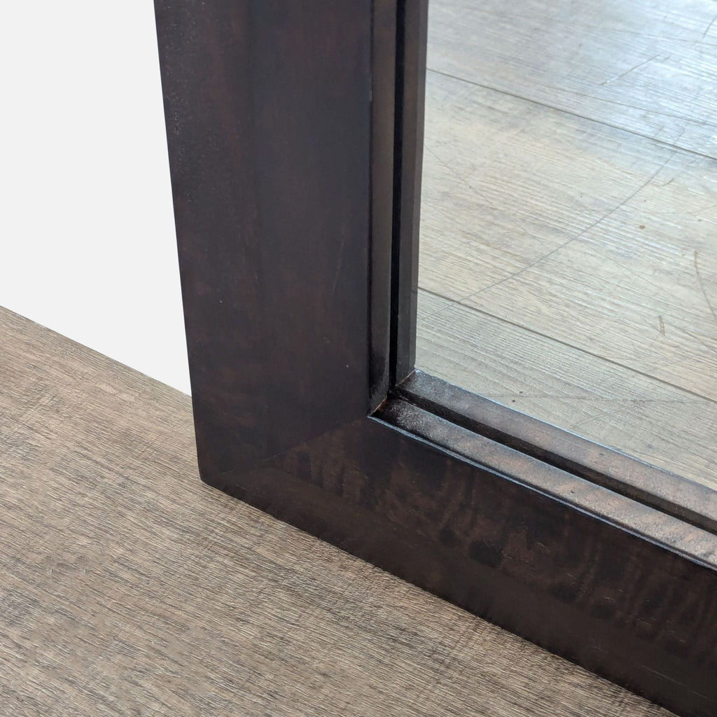 2. Closeup of the bottom corner of a dark wood-framed Crate and Barrel mirror resting on the floor.