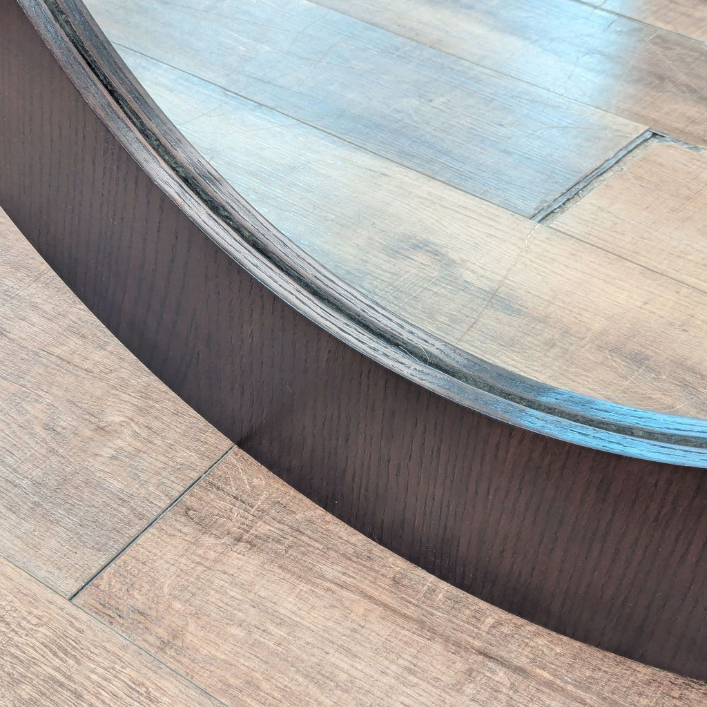 3. Close-up of the dark wooden frame and edge detail of a round Crate and Barrel mirror on wood flooring.