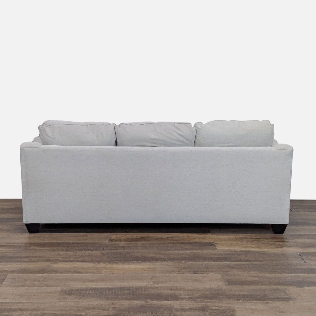 Rear view of a light gray Living Spaces sofa, displaying three back cushions and simple wood feet.