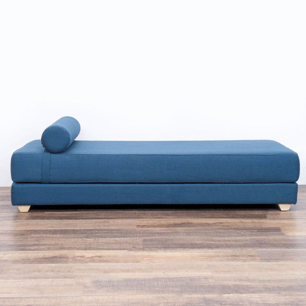 2. Side view of a modern blue CB2 lounge with stacked mattresses and a round bolster cushion, placed on wood flooring.