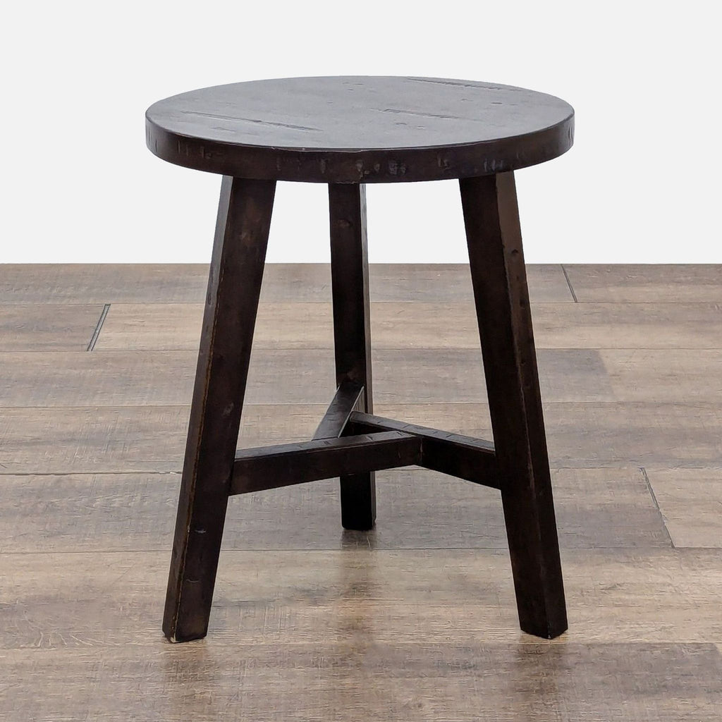 Dark wooden round stool by Reperch with three legs and simple design, placed on a wooden floor.  