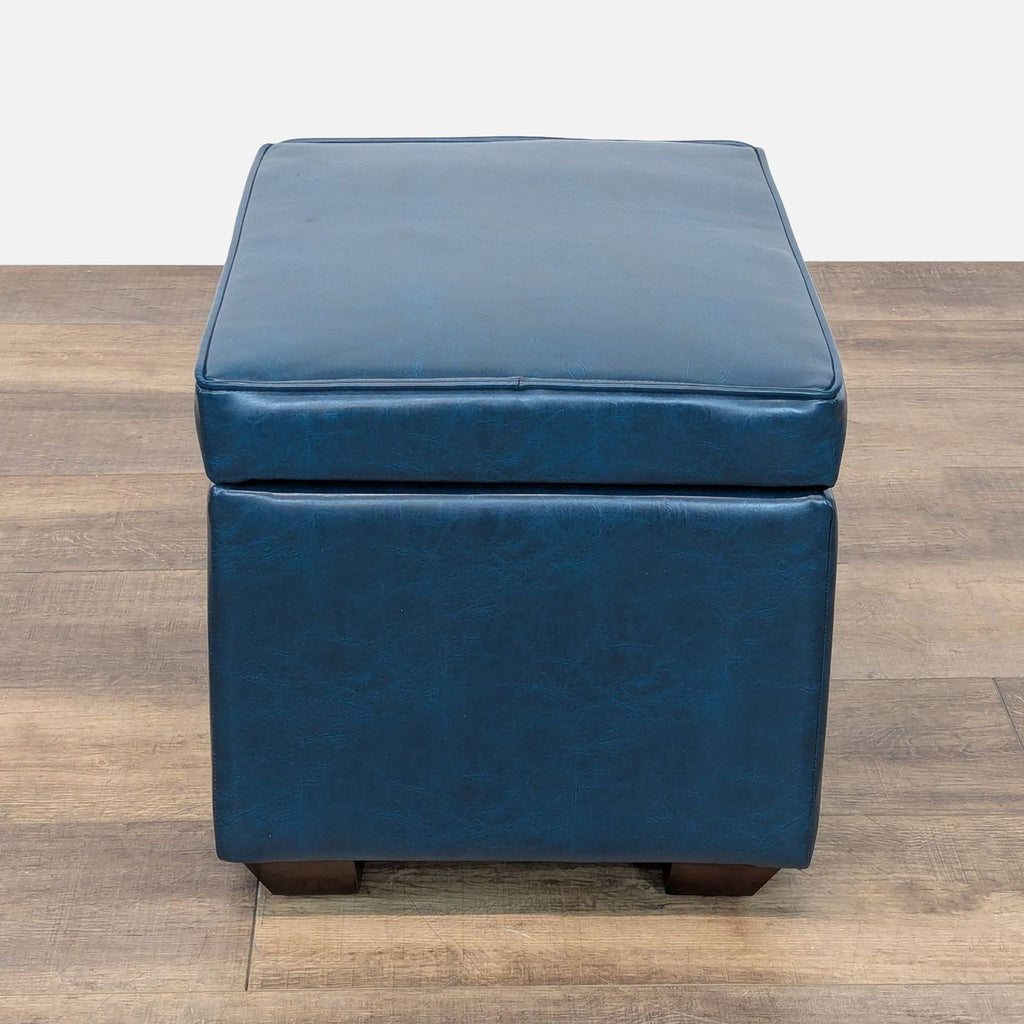 3. Square blue Reperch ottoman with padded seat and wood legs, photographed from the side on wood flooring.