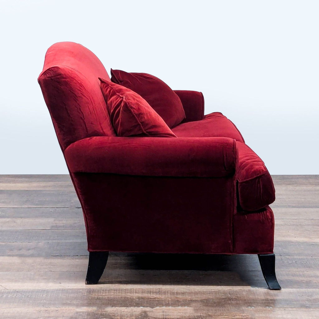2. Side view of red velvet Crate & Barrel loveseat showing rolled arms, plush cushions, and wooden feet, transitional style.