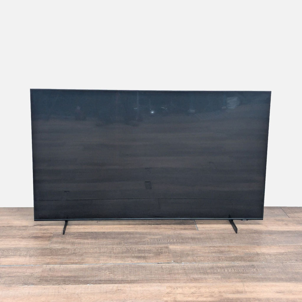 Back view of a Samsung 65-inch TV, showing its large sleek screen and sturdy dual stand on a wood floor.  