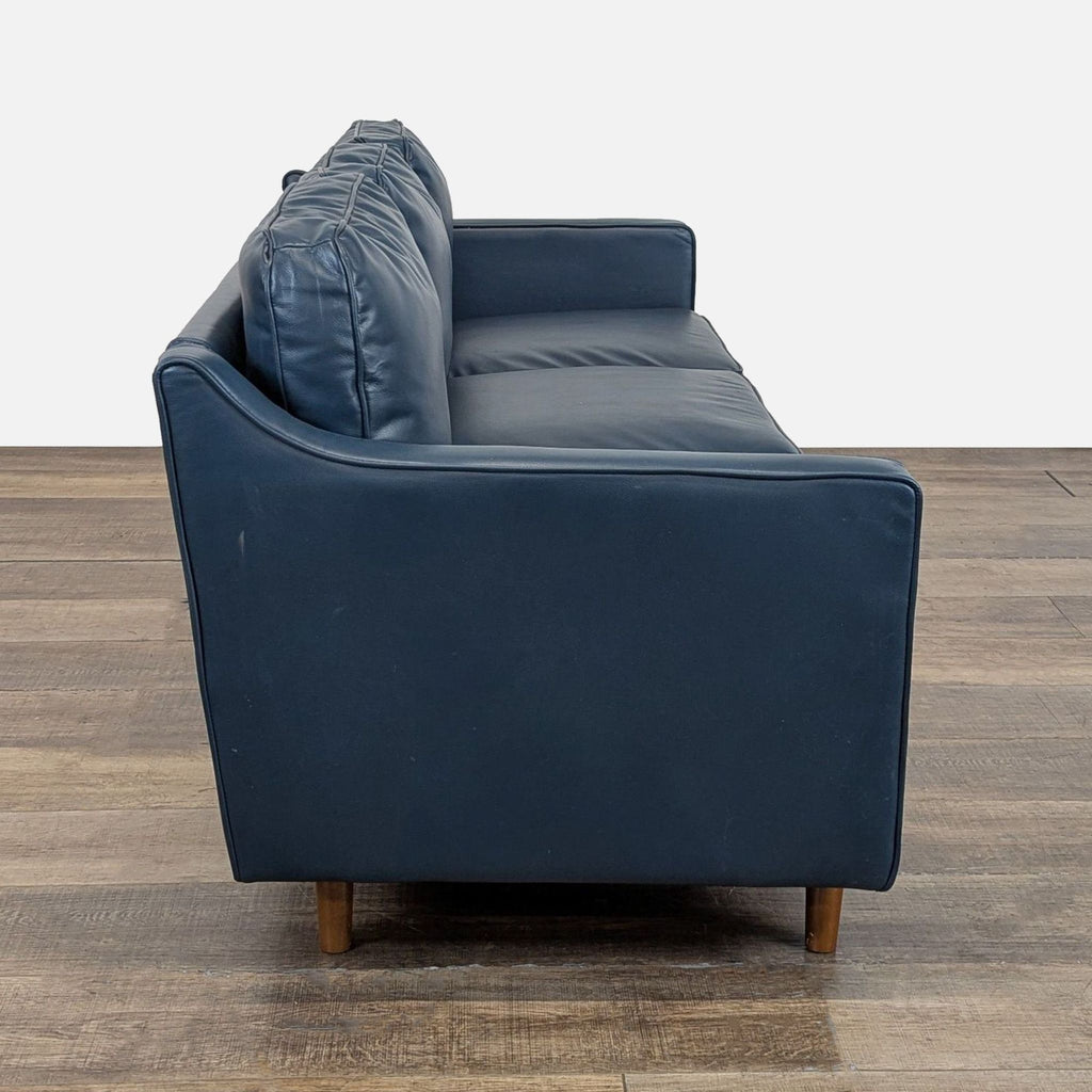 Side profile of a navy faux leather Wayfair 3-seat sofa with wooden legs on a wooden floor.  