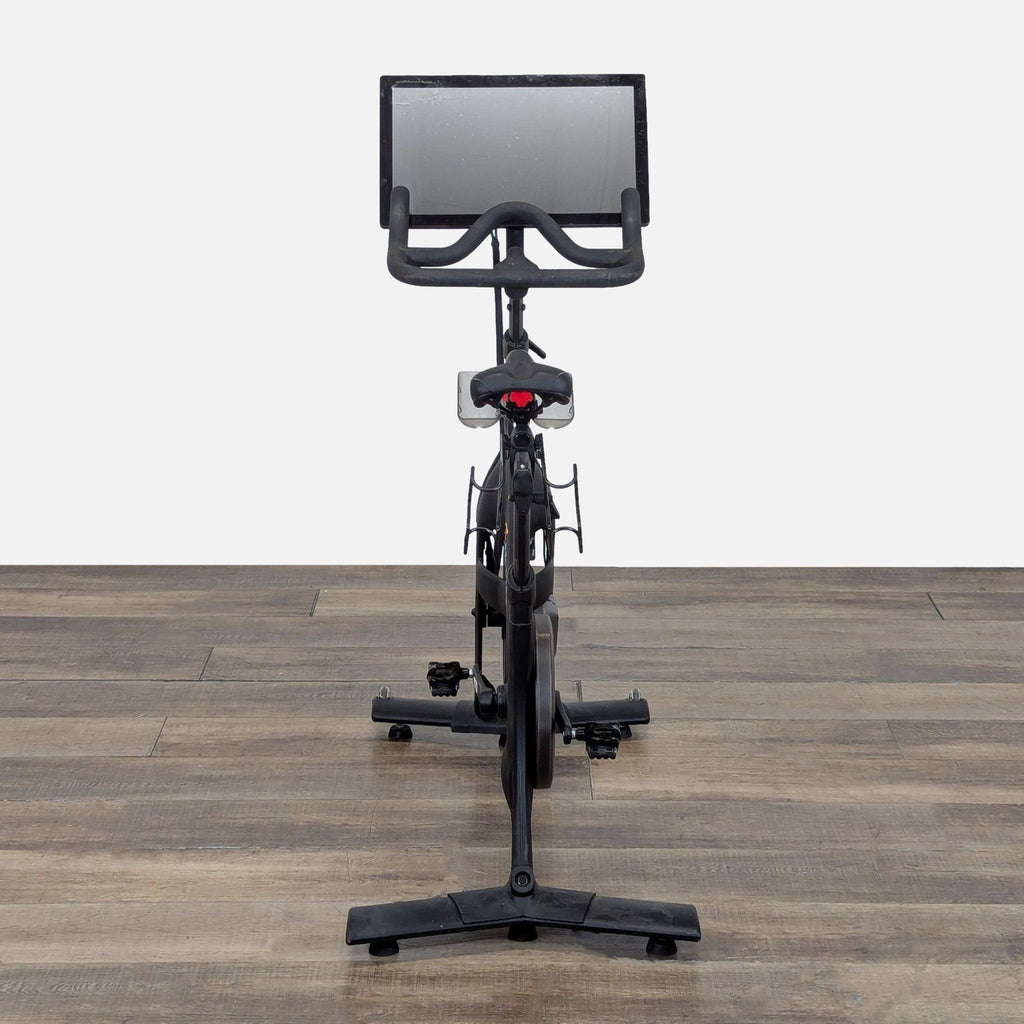 Front view of a Peloton Bike PLTN-RB1VQ with large touchscreen, on a wood floor, for home cardio workouts.  
