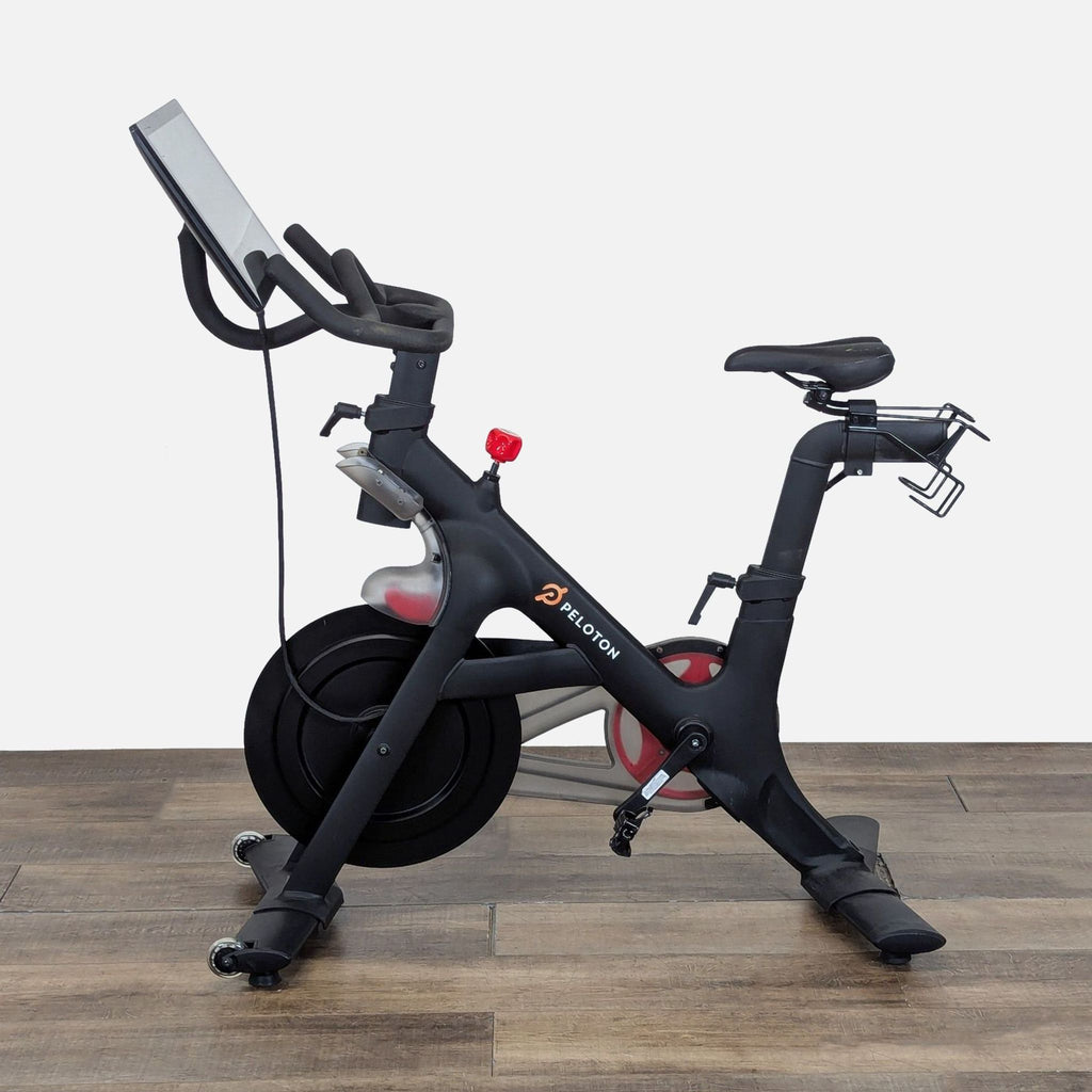 Side view of Peloton exercise bike showing sleek frame, HD touchscreen, and branded details on wooden gym flooring.  