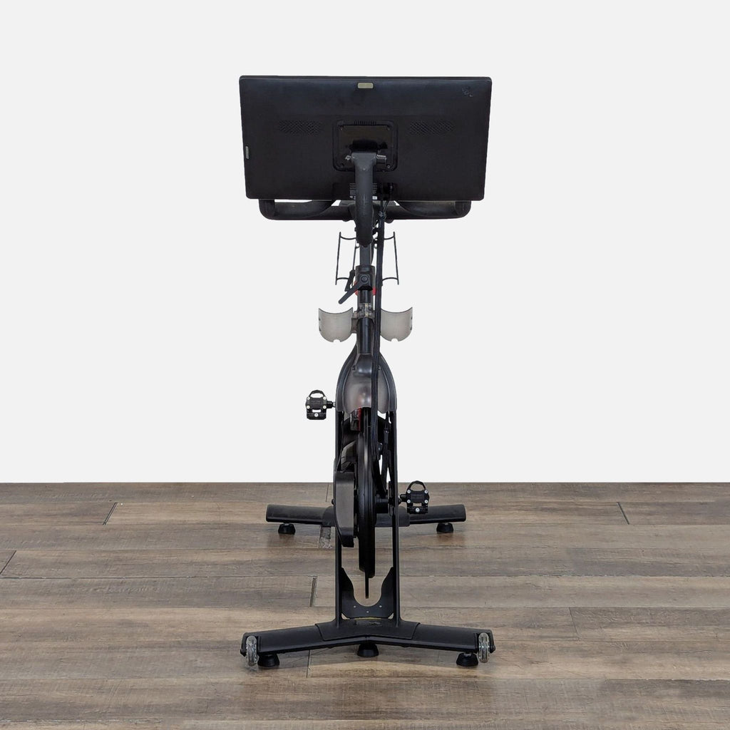 Rear view of Peloton PLTN-RB1VQ indoor bike, displaying adjustable seat and performance screen in a gym setup.