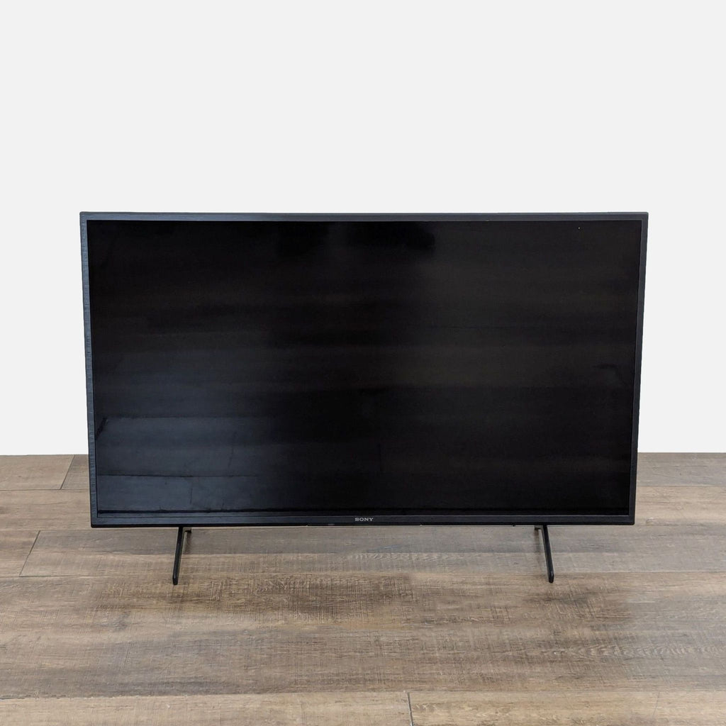 Front view of a Sony KD-43X80J 43-inch 4K Ultra HD Smart TV with a black screen on a wooden floor.  