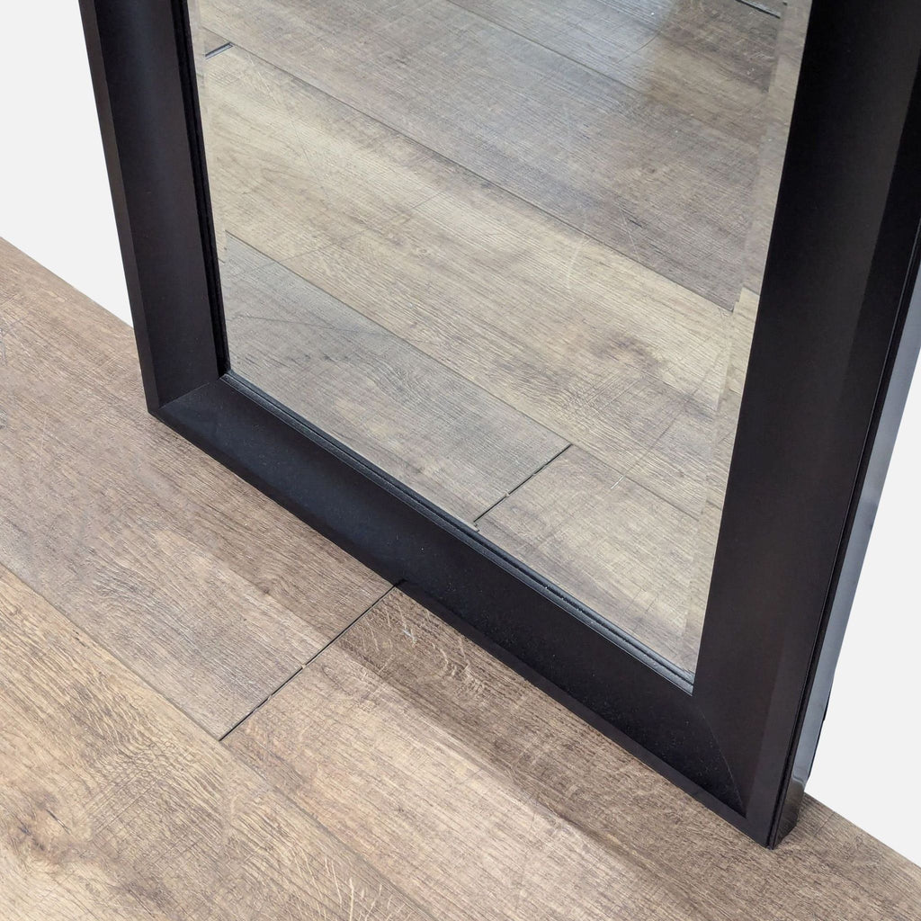 3. Close-up of the bottom corner of a Target rectangular mirror with a sleek black frame on a wood floor.
