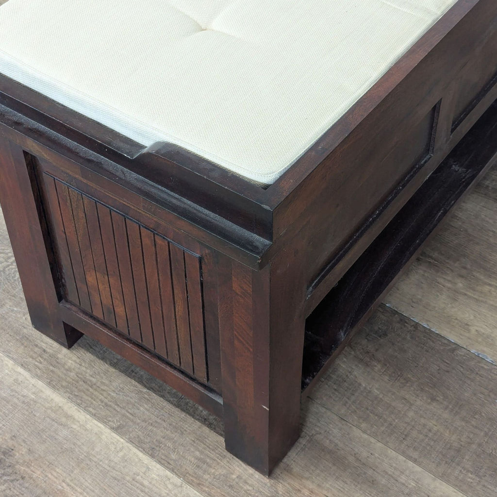 3. Close-up of the side detail on a Pottery Barn wooden bench with a beige cushioned top and paneled end.  