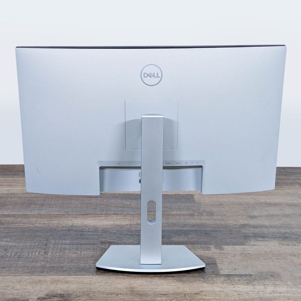 Back view of the Dell S2722QC monitor, showing its silver stand and rear branding on a wood surface.