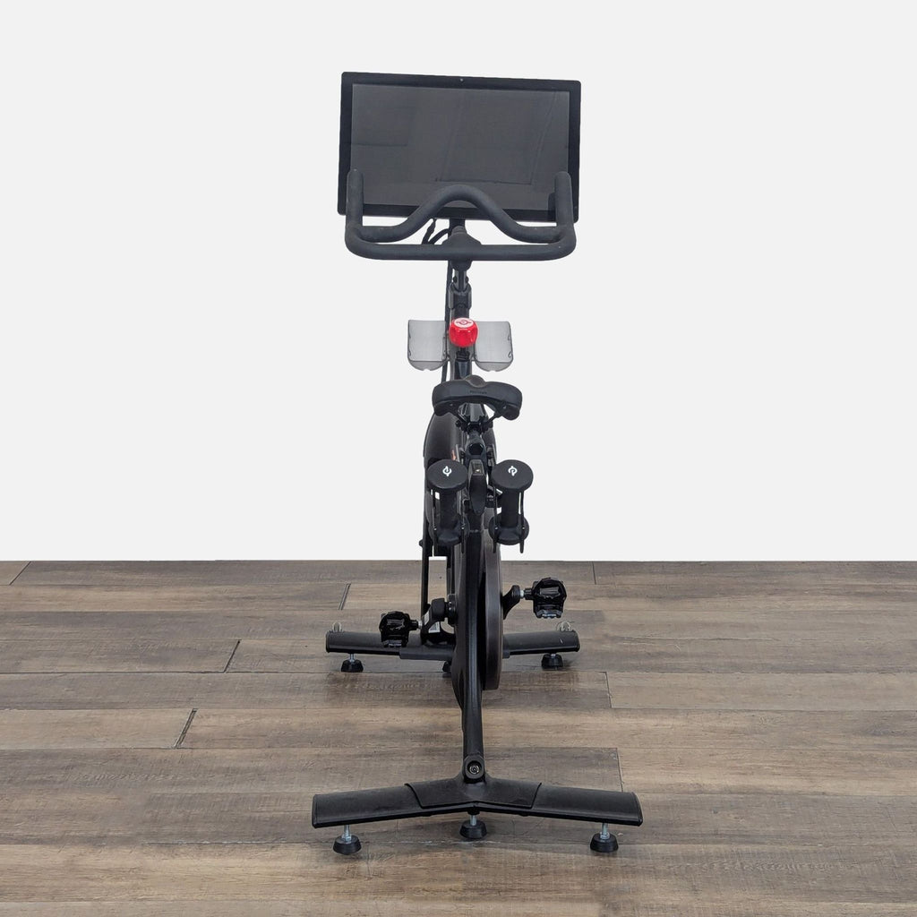 2. Front view of a Peloton gym bike, with a large touchscreen and dual weight holders, set on a wooden floor.