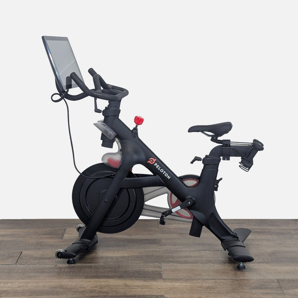 1. Side view of a Peloton exercise bike showing its touchscreen, adjustable seat, and sturdy frame on wood floor.