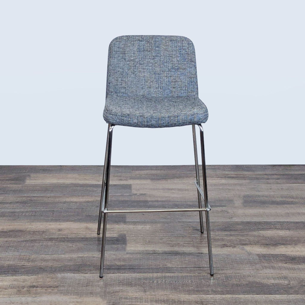 1. Front view of a modern CB2 counter stool with textured blue-gray upholstery and sleek metal legs on a wood floor.