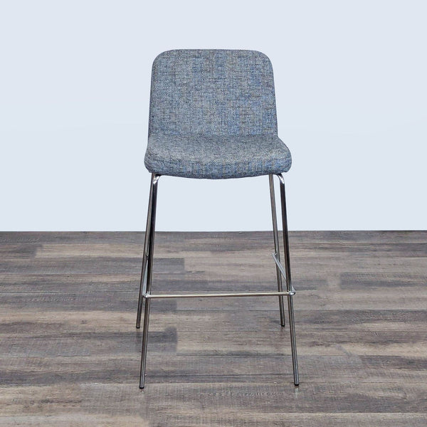 1. Front view of a modern CB2 counter stool with textured blue-gray upholstery and sleek metal legs on a wood floor.