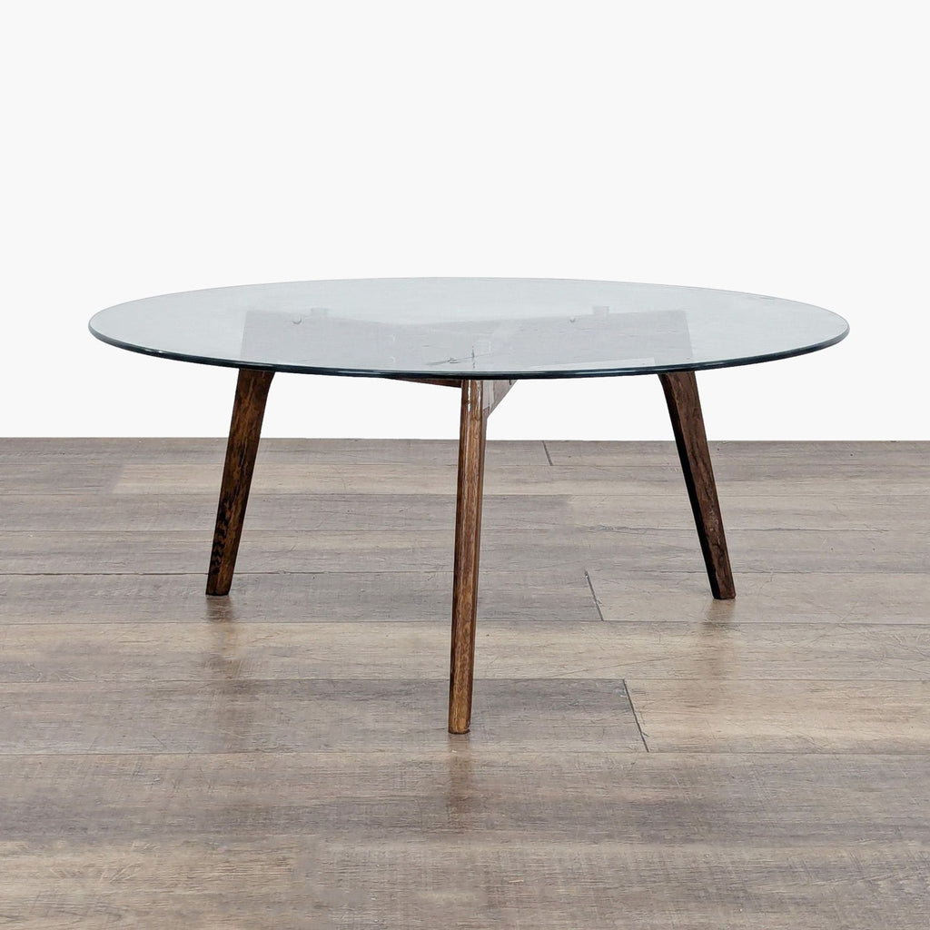 1. Article coffee table with round glass top and three angled wooden legs on a wooden floor.