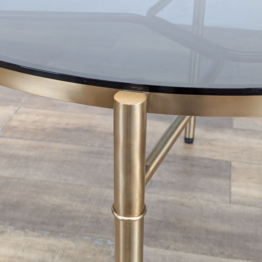 Close-up of the Reperch table’s rounded brass-colored metal base supporting a glass tabletop.  