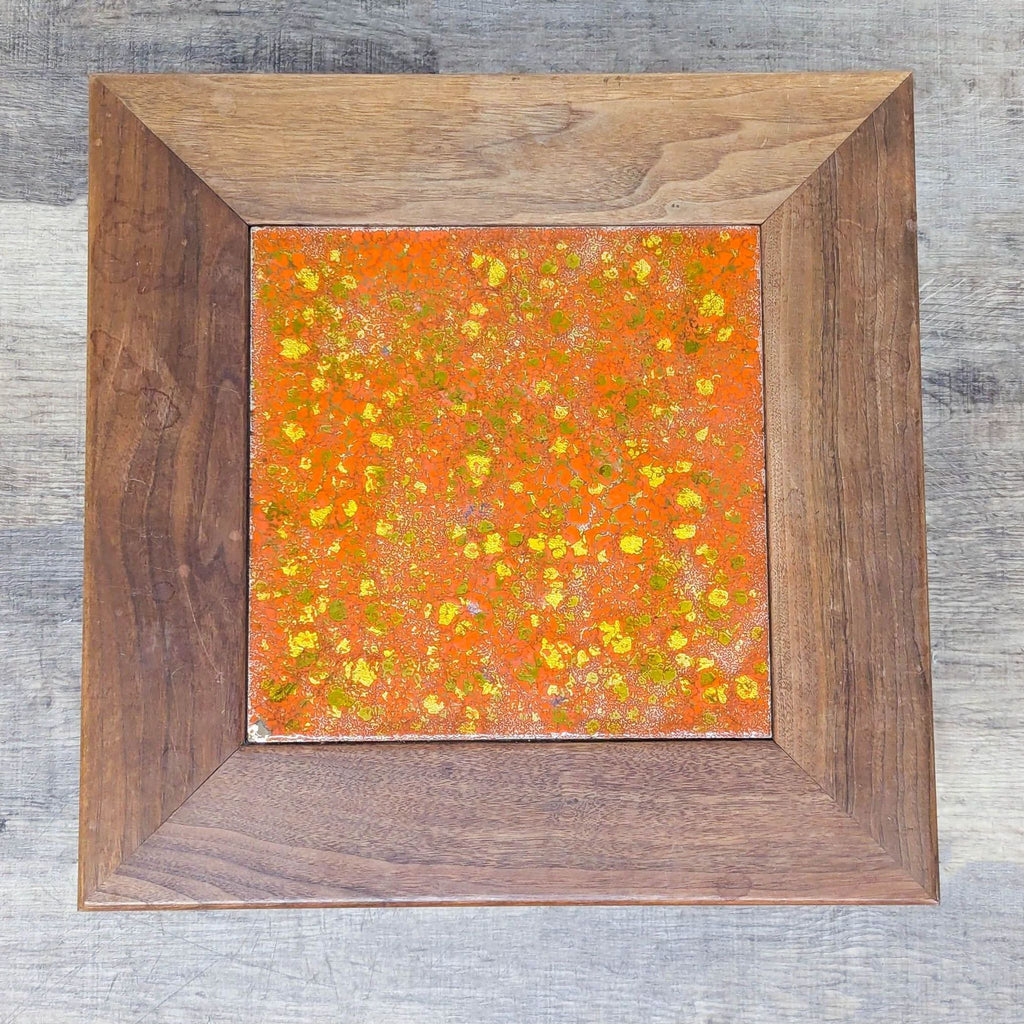 2. Top view of a square table’s surface, showing a mottled orange, yellow, and tan enamel inlay framed by walnut wood.