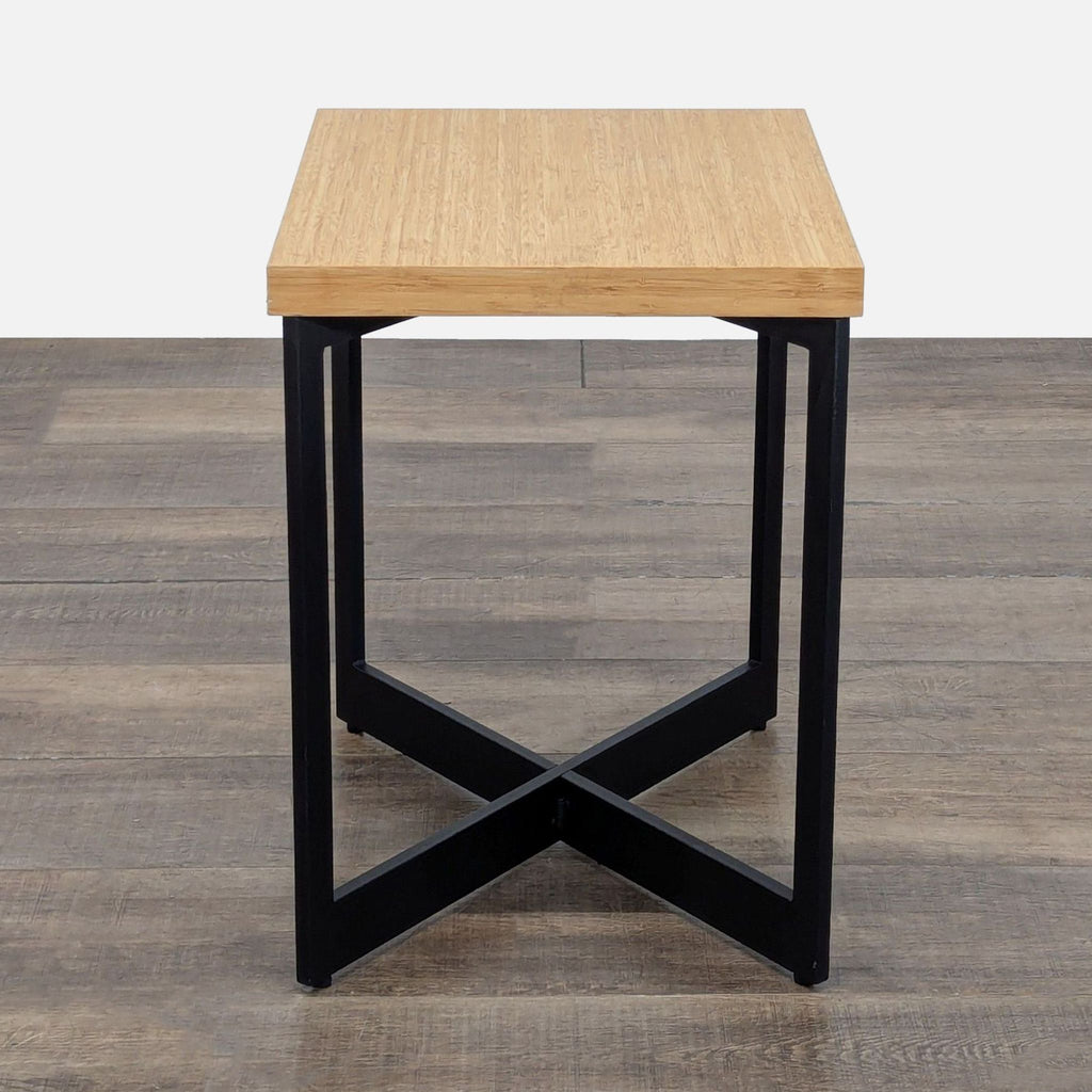 3. Square Reperch end table, wood top and black metal legs with intersecting support, on brown flooring.