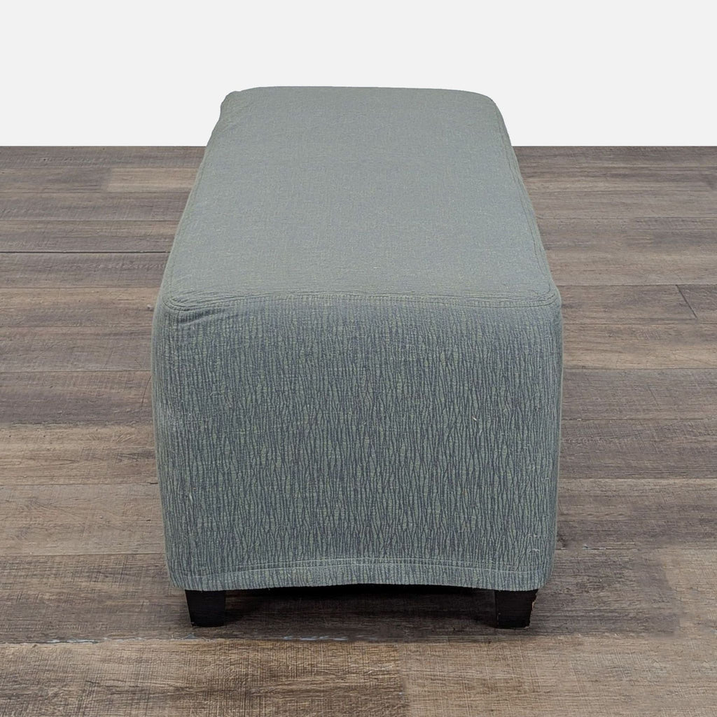 Side view of a modern Reperch ottoman with subtle green upholstery, set on a rustic wooden floor.