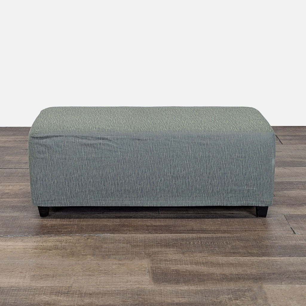 Rectangular Reperch ottoman with muted green fabric cover, shown on a wooden floor, viewed from the front.