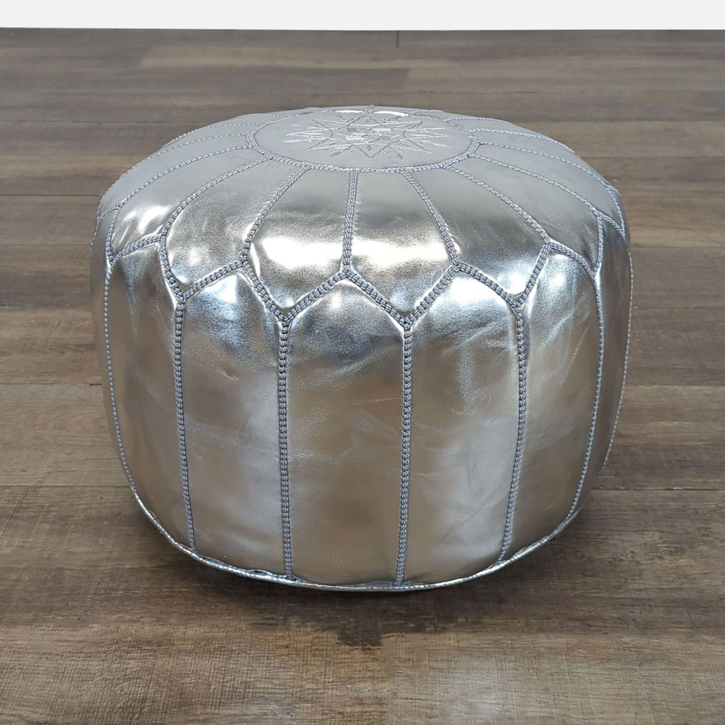 1. Round metallic silver ottoman from Mistana with decorative stitching, shown on a wood floor.
