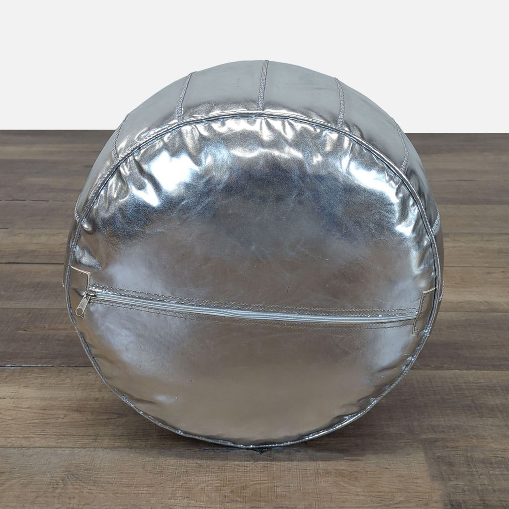 3. Bottom view of Mistana ottoman in reflective silver with a zipper closure, placed on a wood surface.