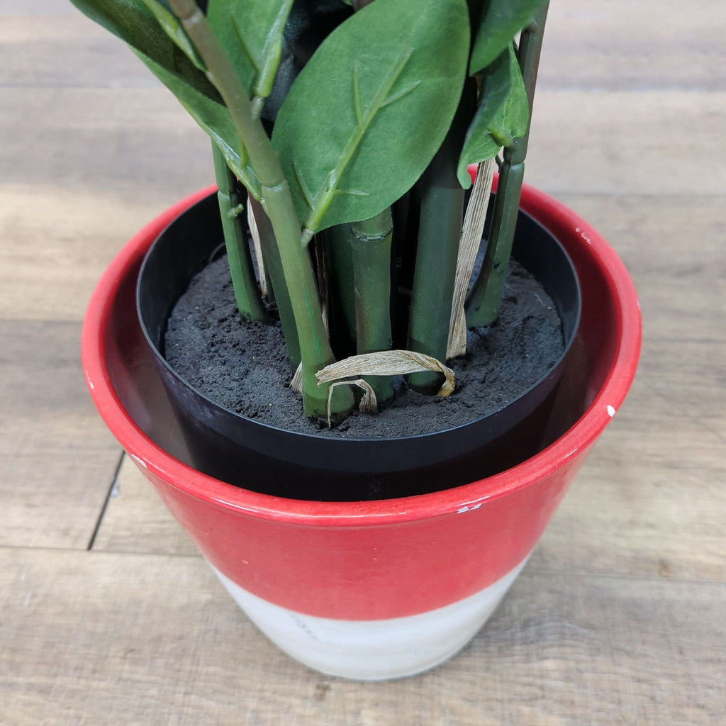 2. Close-up of the base of an artificial plant in black soil, placed inside a red and white Reperch pot.